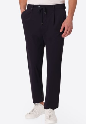 Uomo che indossa pantaloni neri con coulisse e scarpe da ginnastica bianche, in piedi con una mano in tasca su sfondo bianco.