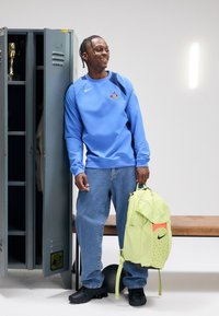 Bluza albastră cu logo, blugi din denim și pantofi negri. Ține un rucsac verde deschis cu un design tipărit și un accent portocaliu.