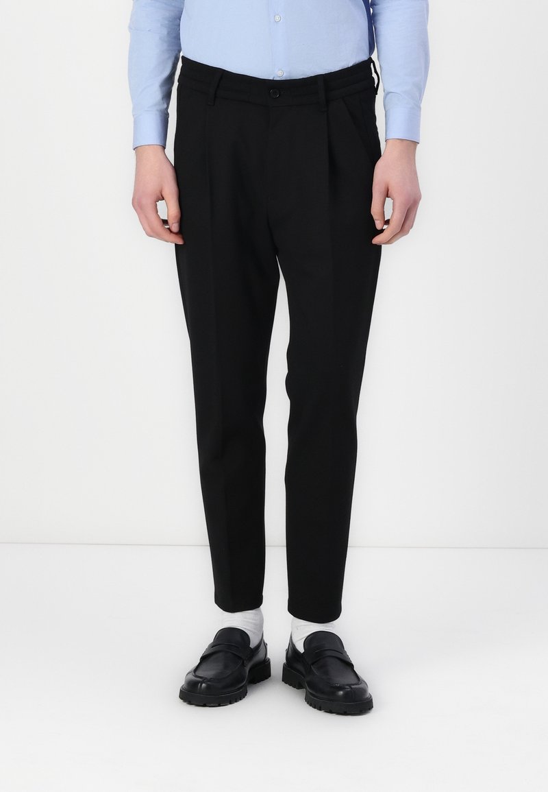 Personne portant un pantalon noir sur mesure, une chemise bleu clair boutonnée, des chaussettes blanches et des mocassins en cuir noir, debout devant un fond blanc uni.