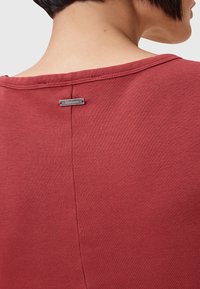 Röd texturerad topp med rundad halsringning och en synlig söm längs ryggen. Silverfärgad märkeslapp fäst nära halsen.