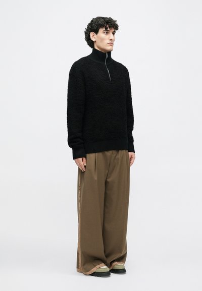 Schwarzer, strukturierter Pullover mit hohem Kragen und einem Viertel-Reißverschluss, kombiniert mit lockeren braunen Hosen und dunklen Schuhen. Schlichtes, modernes Design.