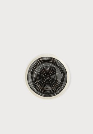 Assiette en céramique avec un motif circulaire de points concentriques noirs et gris foncé sur fond blanc, centrée sur une surface claire et unie.