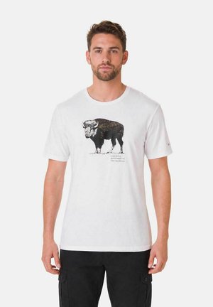 Mann mit kurzen braunen Haaren, der ein weißes T-Shirt mit schwarzer Büffel-Grafik und schwarzen Cargohosen trägt, steht vor einem einfarbigen Hintergrund.