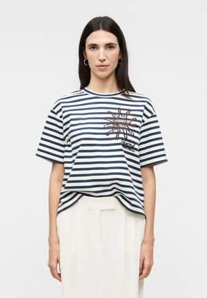 Γυναίκα με μακριά σκούρα μαλλιά φοράει μπλε ναυτικό και λευκό ριγέ μπλουζάκι με καφέ σχέδιο ήλιου και λευκό ψηλόμεσο παντελόνι μπροστά σε απλό φόντο.