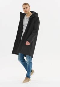 Svart lång regnkappa med huva, med knappslå och en vadderad insida, matchad med ljusblå jeans och beiga sneakers.