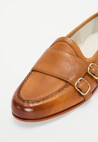 Mocassino in pelle marrone con una superficie liscia, dotato di una fascia con due fibbie dorate, punta arrotondata e cuciture decorative.