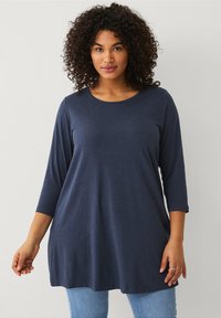 Marineblauw lang straat shirt met een ronde halslijn, zachte stof, losse pasvorm en een zoom die onder de heupen uitkomt; gedragen met lichtblauwe jeans.