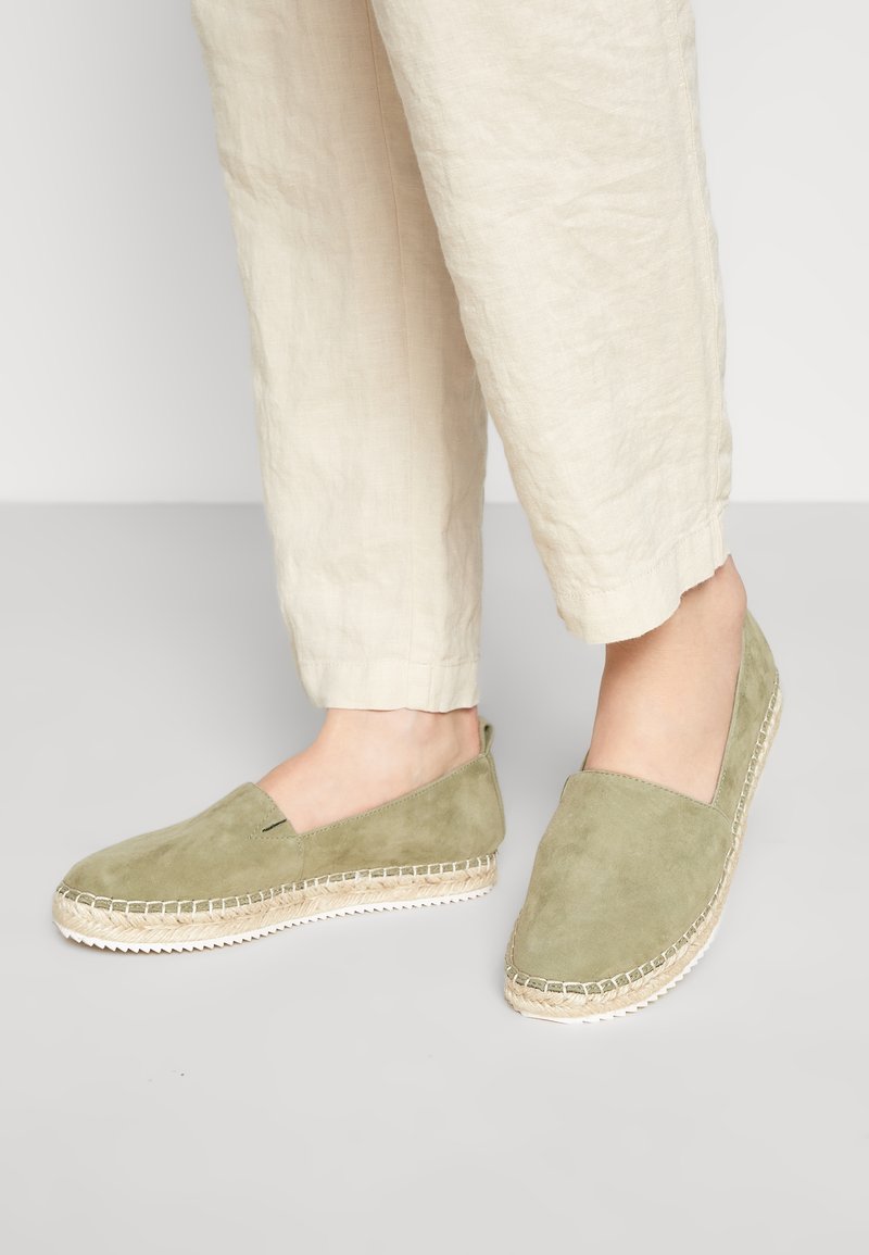 Espadrilles en daim vert avec une semelle en jute et des détails cousus, dotées d'un bout et de côtés fermés, associées à un pantalon en lin beige.
