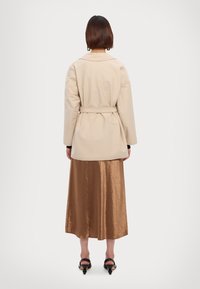 Beige Jacke mit Gürtel in lockerer Passform, kombiniert mit einem glänzenden, knielangen braunen Rock. Glatte Textur, kombiniert mit schlichten Schuhen.