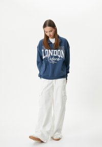 Sudadera azul marino con gráfico en blanco "LONDON Oxford", cuello con cremallera; combinada con pantalones cargo blancos de corte holgado y zapatos beige.