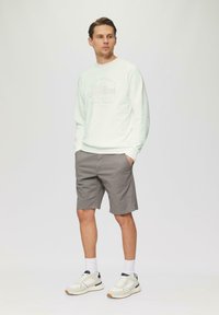 Felpa grigia chiara con stampa, abbinata a pantaloni corti grigi. Il modello indossa sneakers bianche e calzini corti, in piedi contro uno sfondo neutro.