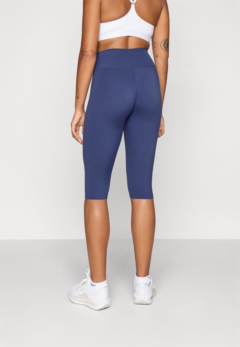 Femme portant des leggings courts taille haute bleu marine, un soutien-gorge de sport blanc, des chaussettes blanches et des chaussures de sport blanches, debout de dos sur un fond uni.