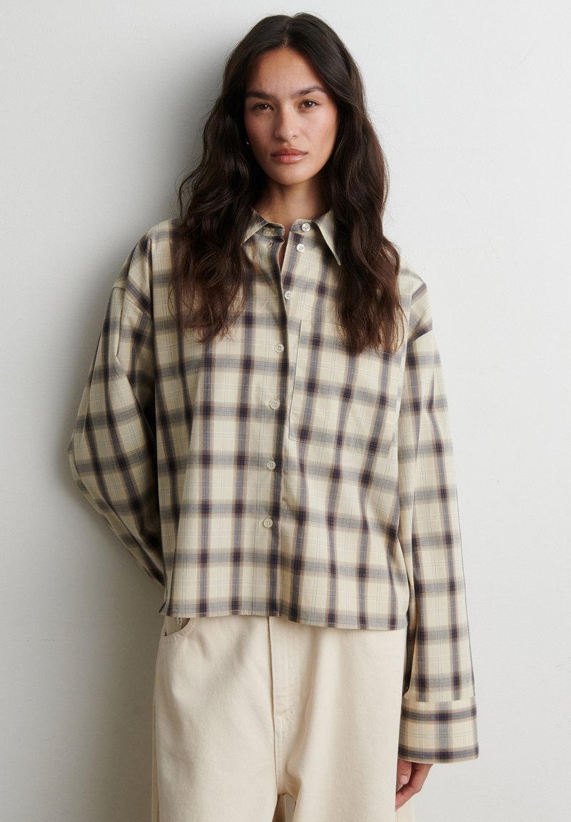 Femme aux longs cheveux foncés portant une chemise à carreaux beige et grise boutonnée et un pantalon beige clair, debout devant un mur blanc uni.