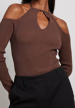 Femme portant un pull marron côtelé à épaules dénudées avec un col à découpe circulaire, associé à un pantalon taille haute noir.