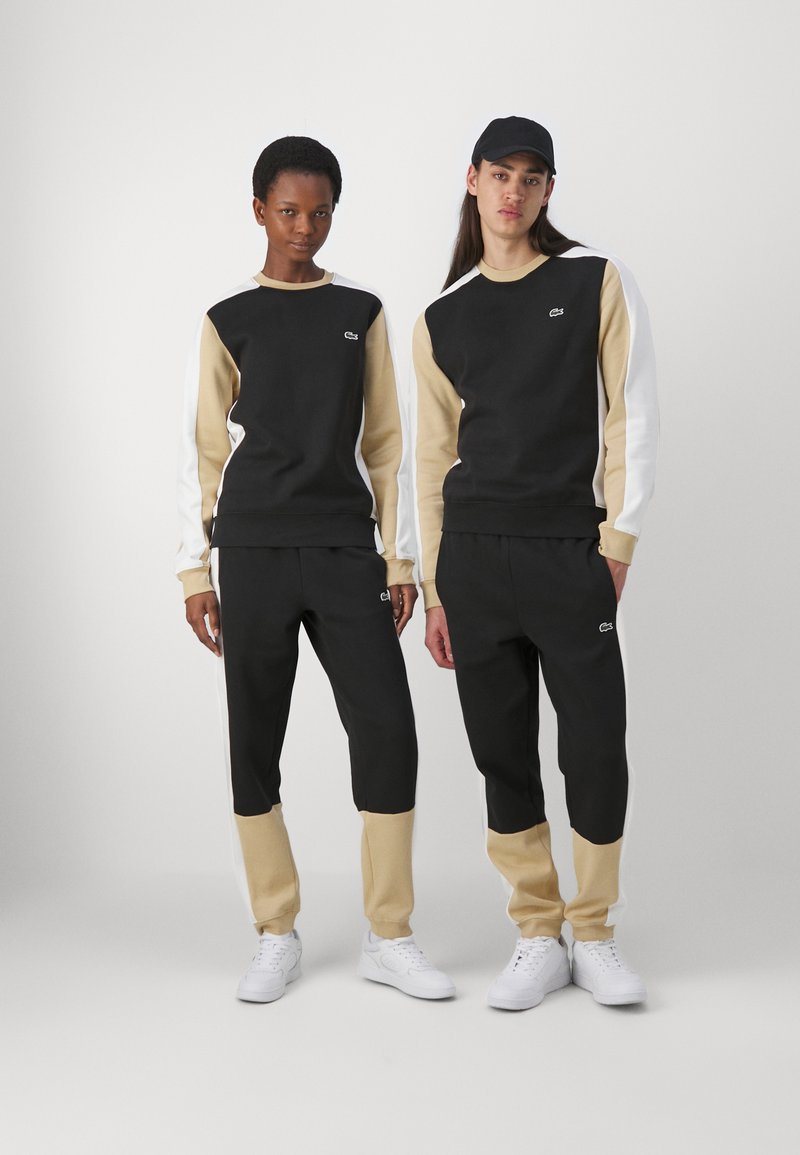 Ensemble de vêtements de sport noir et beige avec un design en blocs de couleur. Les sweat-shirts ont des poignets côtelés ; les pantalons possèdent des chevilles élastiquées ; détail du logo sur la poitrine.