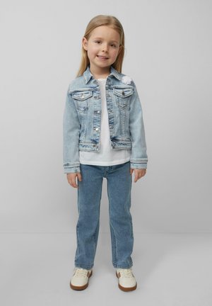 Veste en denim bleu clair avec des boutons argentés, deux poches poitrine et un accent floral blanc. Portée sur une chemise blanche et un jean bleu clair.