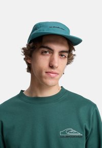 Gorra color teal hecha de un tejido ligero con visera plana, que presenta el texto "Quiksilver" en negro. Se lleva con una sudadera verde oscura con detalle de logo.