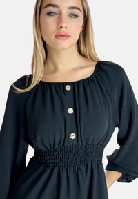 Schwarzes Kleid mit quadratischem Ausschnitt, geraffter Taille und langen Ärmeln, ausgestattet mit fünf cremefarbenen Knöpfen auf der Vorderseite. Glatte Stoffstruktur.
