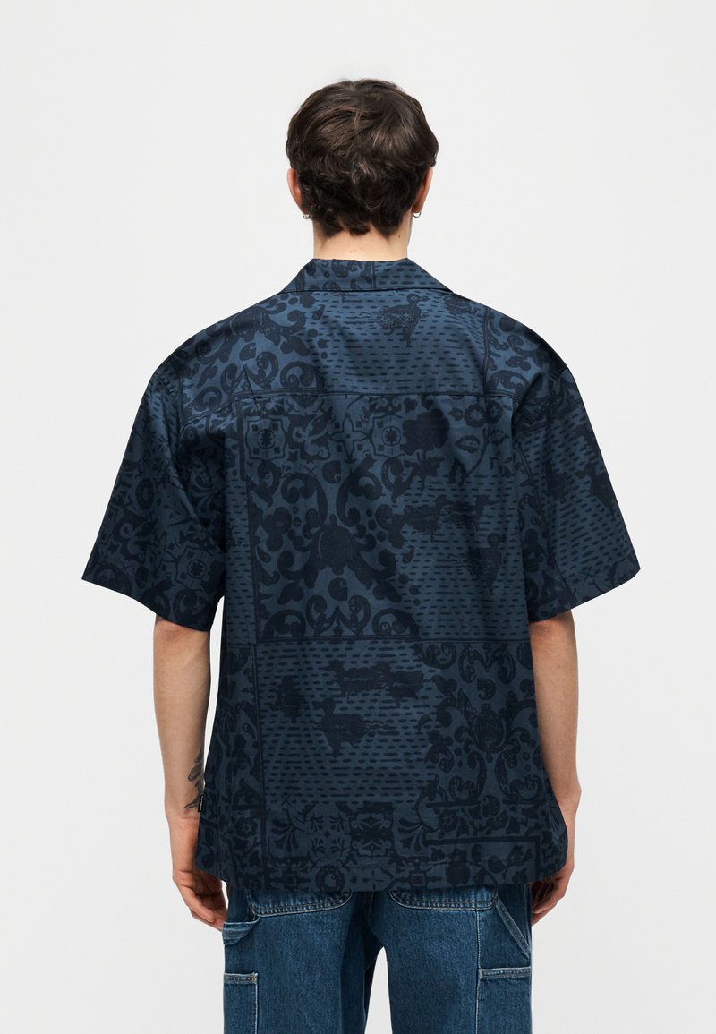 Persona che indossa una camicia a maniche corte blu scuro con un motivo floreale e geometrico sottile, abbinata a jeans blu, vista da dietro.