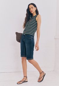 Top fără mâneci cu dungi verzi și albe asortat cu pantaloni scurți din denim închis, întorși la tiv. Modelul poartă o geantă plic maro împletită. Sandalele simple completează ținuta.