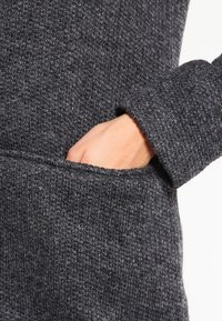 Szary sweter dzianinowy o strukturalnym wykończeniu, z ręką sięgającą do bocznej kieszeni, widocznymi szwami i detalami mankietów.