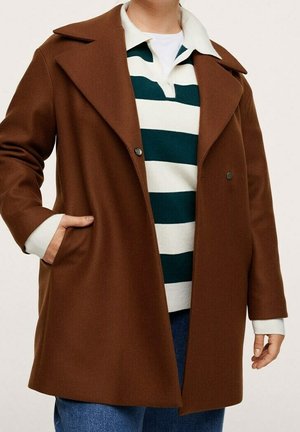 Personne portant un manteau marron croisé sur une chemise blanche à rayures vert foncé avec col et un jean bleu, une main dans la poche.