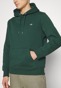 Dark green hoodie made of soft cotton blend, featuring a kangaroo pocket, adjustable drawstring hood, and small embroidered logo on the chest.