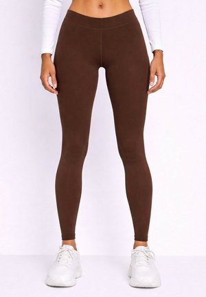 Femme portant un legging marron taille haute, un crop top blanc à manches longues et des chaussures de sport blanches, debout devant un fond uni.