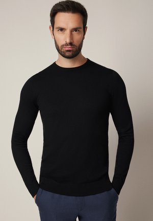 Czarny sweter z długim rękawem wykonany z miękkiego dzianinowego materiału, z okrągłym dekoltem oraz ściągaczami na mankietach i dole. Dopasowany fason o gładkiej fakturze.