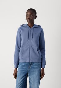 Sweat à capuche zippé bleu avec un logo sur la poitrine, deux poches avant et des poignets côtelés. Porté avec un jean bleu clair.