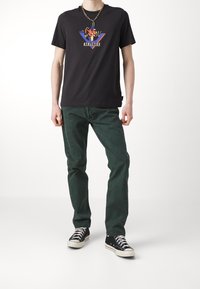 Person in einem schwarzen Grafik-T-Shirt, dunklen grünen Jeans, schwarzen High-Top-Sneakern und einer goldenen Kettenhalskette vor einem weißen Hintergrund.
