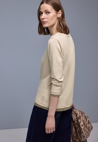 Beige long-sleeve shirt with a textured brown trim at the hem and cuffs, made of soft fabric, paired with dark denim pants and a brown shawl.