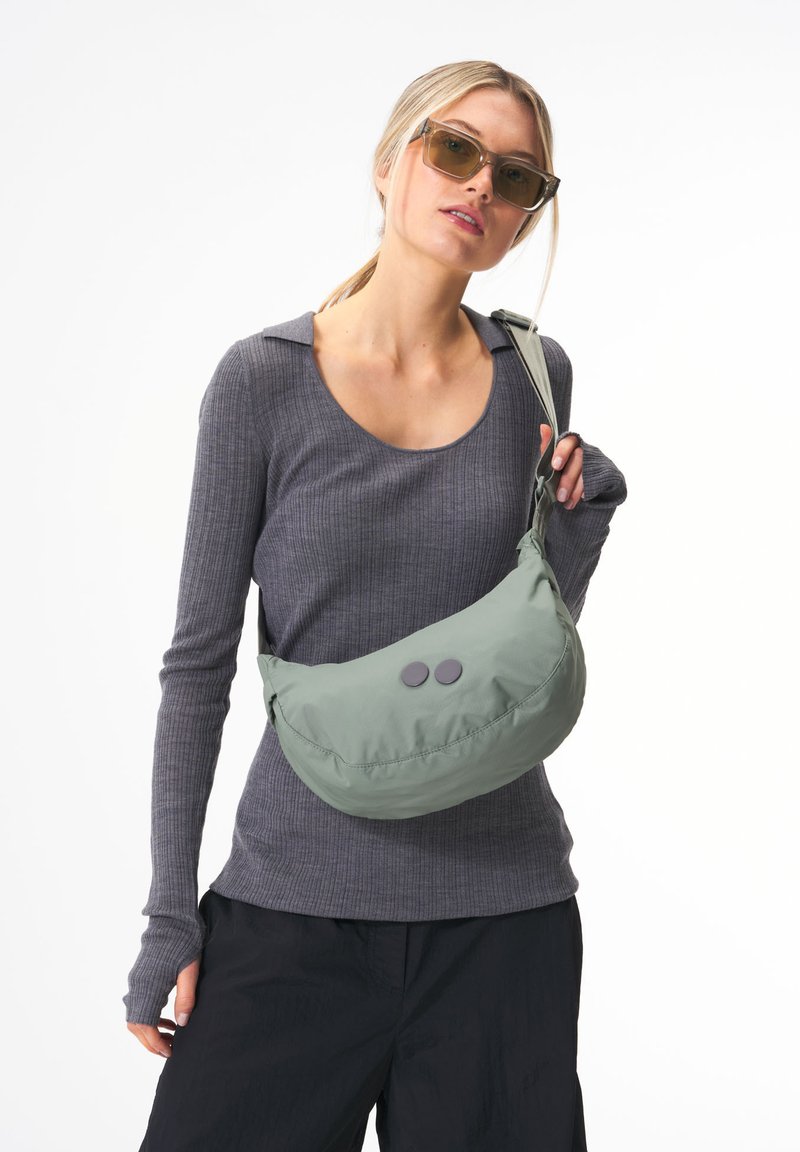 Grüne Nylontasche mit Schultergurt in runder Form, zwei kreisförmige graue Knöpfe und verstellbarem Riemen, getragen über einem grauen, gerippten Langarmoberteil.