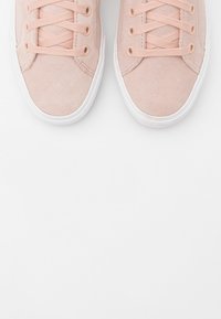 Světle růžové semišové tenisky s bílými gumovými podrážkami, vybavené kulatými průchody na tkaničky a plochými tkaničkami. Hladká textura s minimalistickým designem.