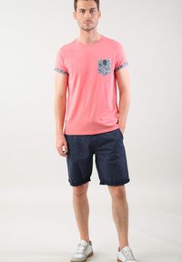 Camiseta de hombre color coral con bolsillo estampado y pantalones cortos azul marino con dobladillo. Tejido ligero, corte casual, combinada con zapatillas blancas.