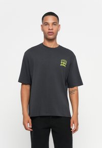 Moški nosi ohlapen črn T-shirt Adidas z majhnim rumenim grafičnim motivom na prsih in črne hlače, stoji ob enobarvni beli ozadju.