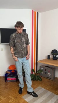 Camouflage short-sleeve shirt with light gray and brown patterns, paired with light blue jeans and black sneakers, standing indoors.