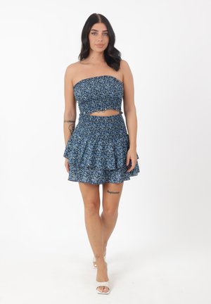 Joven mujer con top corto sin tirantes azul con flores y falda mini de volantes, caminando hacia adelante sobre fondo blanco.