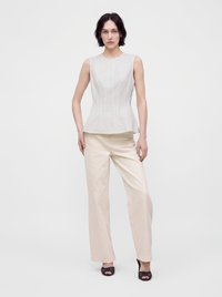 Femme aux cheveux courts et foncés, vêtue d'un haut blanc sans manches, d'un pantalon ample couleur crème et de talons noirs ouverts, debout devant un fond uni.