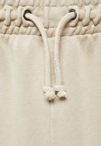 Beige puuvillaiset collegehousut, joissa on ribbireunusvyö, säädettävät nyörit solmittuine päineen ja sileä tekstuurinen pinta.