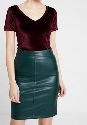 Mujer con una blusa de terciopelo burdeos de manga corta metida dentro de una falda lápiz de cuero verde oscuro de cintura alta.