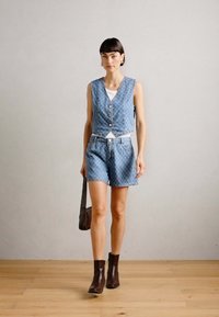 Denim prsluk s dugmadi, u kombinaciji s odgovarajućim traper šortsevima s dijamantnim uzorkom. Model nosi smeđe gležnjače i nosi torbu na rame.