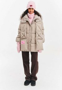 Světlá béžová péřová bunda s diamantovým prošitým vzorem, náprsními kapsami na knoflíky, růžovou nášivkou, doplněná růžovými rukavicemi, černými kalhotami a černými botami.