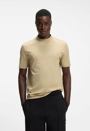 Jeune homme aux cheveux courts porte un t-shirt beige à manches courtes à col montant et un pantalon noir, debout les mains dans les poches sur fond blanc.