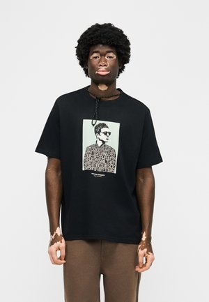 Νέος άνδρας με λεύκη φοράει μαύρο μπλουζάκι με μονοχρωματικό πορτρέτο και κείμενο, στέκεται μπροστά σε ένα απλό ανοιχτό φόντο.