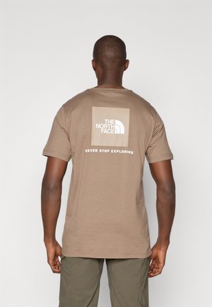 Man draagt beige The North Face T-shirt met logo en de tekst "Never Stop Exploring" op de achterkant, gecombineerd met olijfgroene broek.