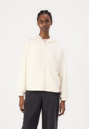 Sudadera cremosa con cremallera y capucha, bolsillo canguro y puños acanalados. Combinada con pantalones gris oscuro, la tela tiene un acabado suave y texturizado.