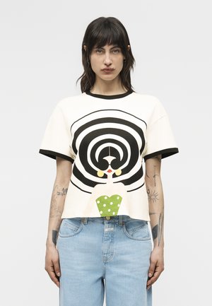 Γυναίκα με σκούρα μαλλιά που φοράει λευκό μπλουζάκι με μαύρους ομόκεντρους κύκλους και μια στυλιζαρισμένη γυναικεία φιγούρα σε πράσινο πουά φόρεμα, συνδυασμένο με ανοιχτό μπλε τζιν.