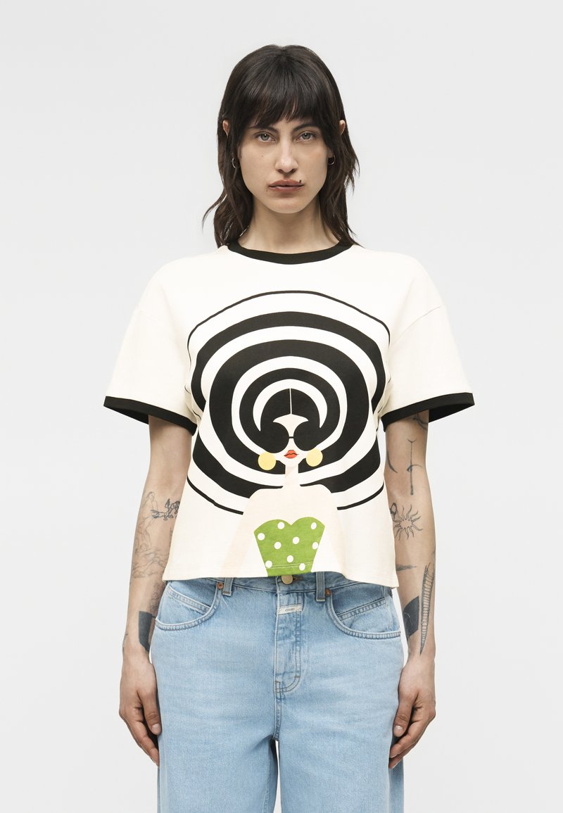 Femeie cu păr închis la culoare, purtând un tricou alb cu cercuri concentrice negre și o figură stilizată de femeie îmbrăcată într-o rochie verde cu buline, asortată cu o pereche de blugi albastru deschis.