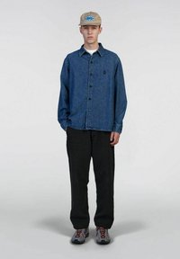 Camicia in denim con bottoni e colletto, abbinata a pantaloni scuri e sneakers. Cappello con logo circolare blu su sfondo beige.